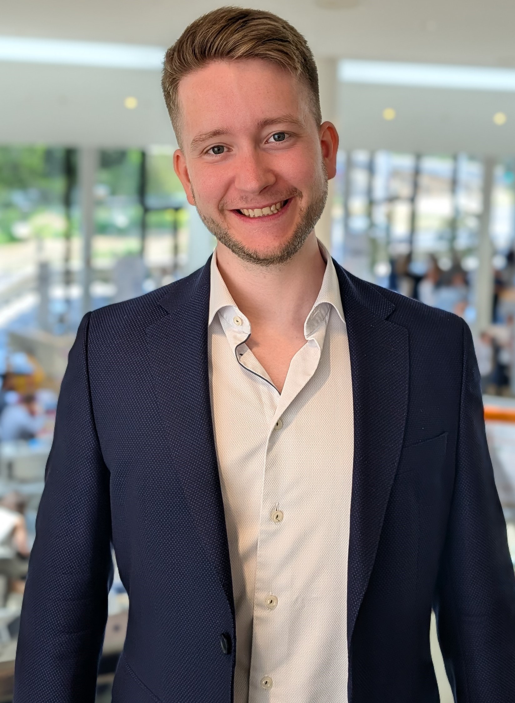 The image shows a person in a navy blazer and light dress shirt standing indoors, likely in a professional or formal setting. The face is obscured by a solid rectangle, so I can’t identify the person.

If you want, I can also help with:
- describing the outfit in more detail
- assessing the photo’s composition or lighting
- suggesting edits for a professional headshot
