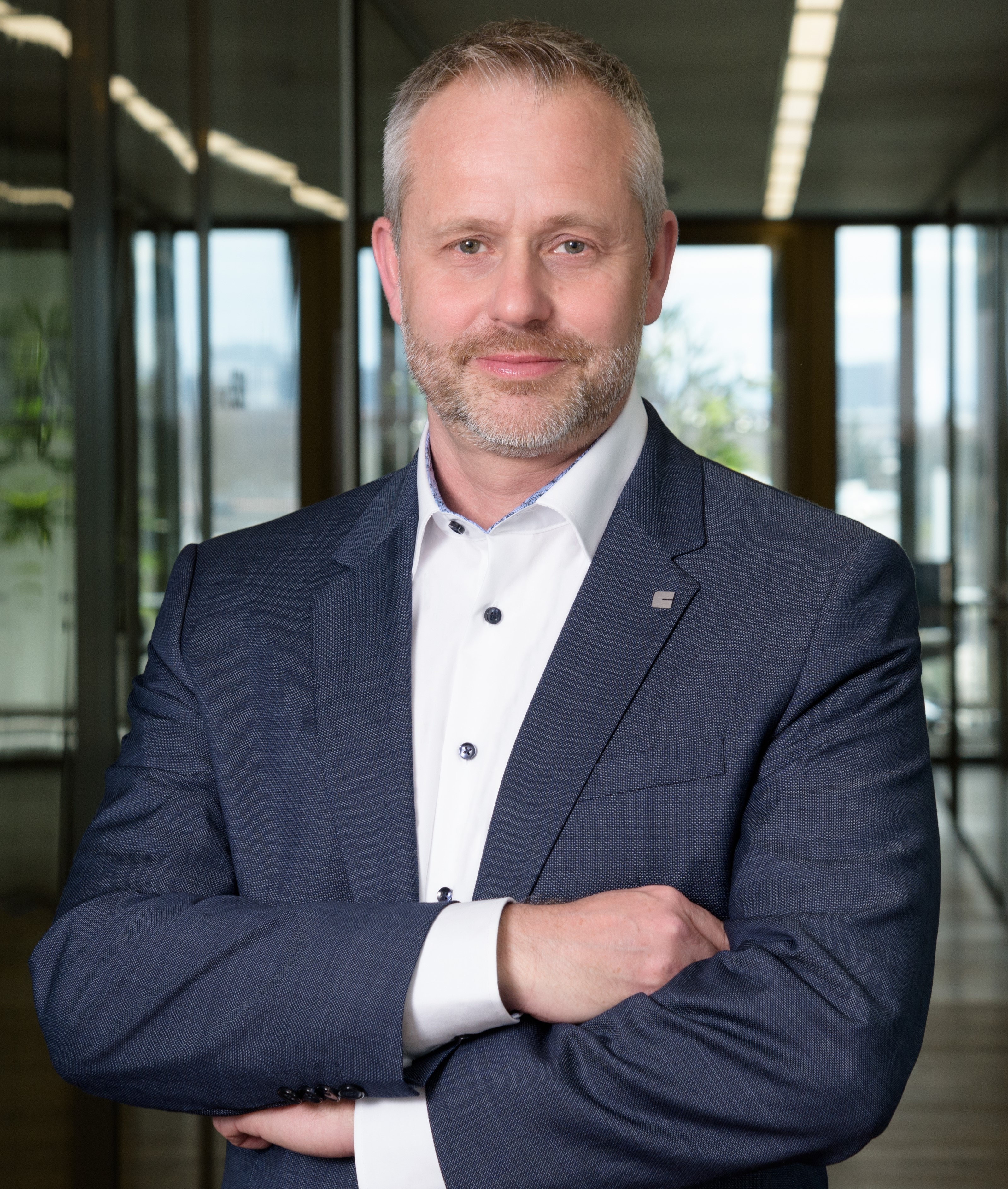 The image shows a man in a business suit standing with his arms crossed in an office-like setting. His face is obscured by a solid rectangular blur. The background has glass walls, reflections, and bright windows, giving it a modern corporate feel.