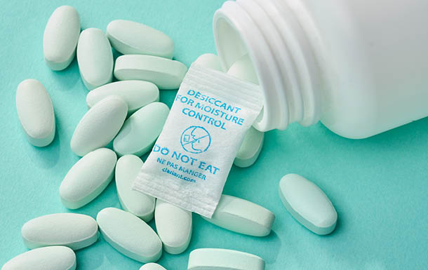 The image shows a white bottle tipped over with white oval pills spilled out onto a light blue surface. Among the pills, there is a small packet labeled "DESICCANT FOR MOISTURE CONTROL" with a warning "DO NOT EAT" written on it. The packet is typically included in medication bottles to absorb moisture and keep the pills dry.