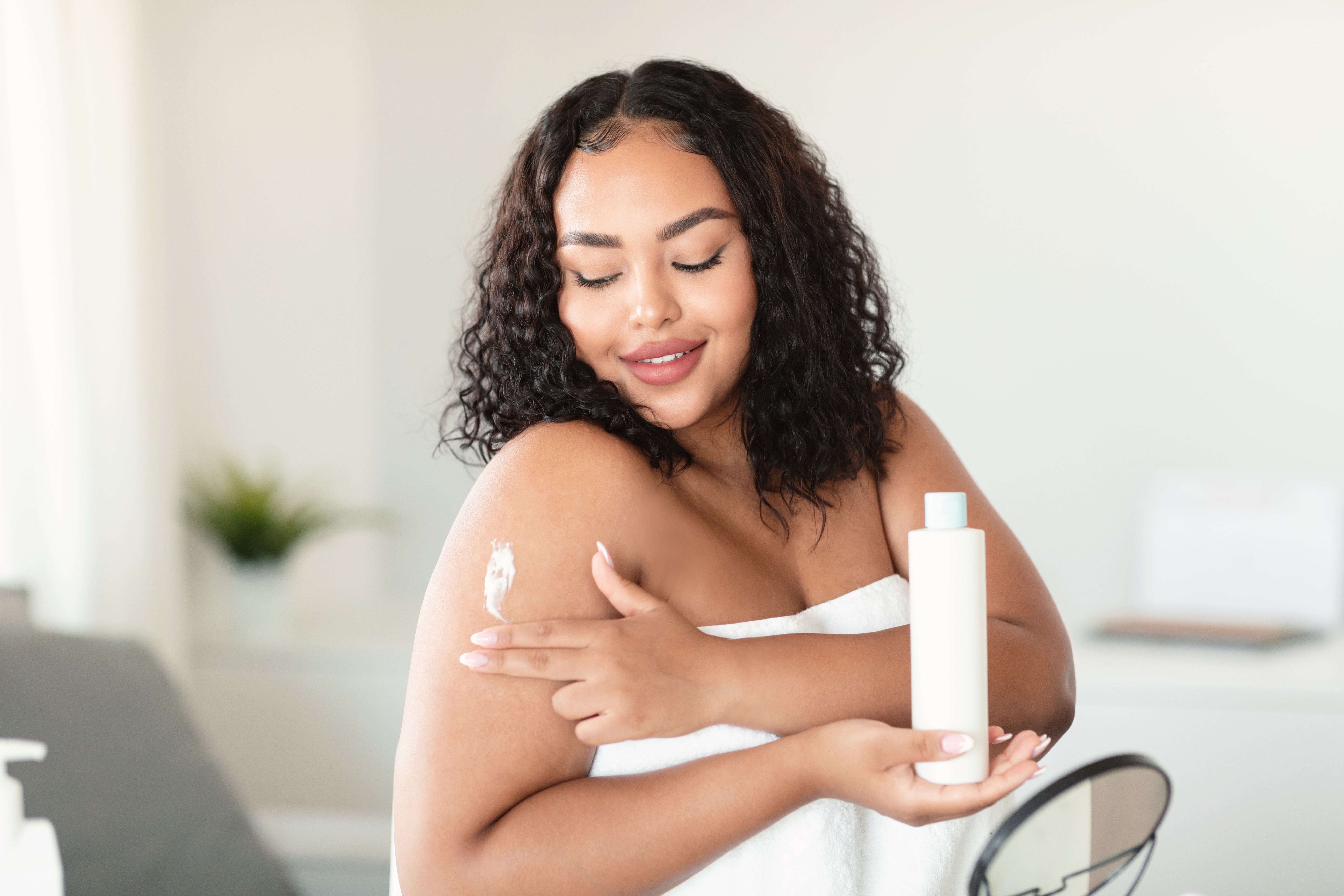 ClariantLovely african american chubby woman wrapped in towel applying body cream on her shoulder af