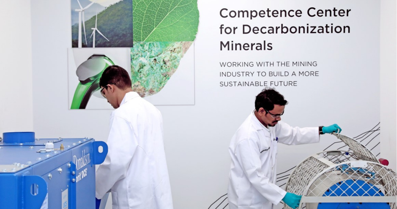 The image shows two individuals wearing white lab coats and blue gloves working with machinery in a laboratory or industrial setting. On the wall behind them, there is text that reads:

"Competence Center for Decarbonization Minerals
WORKING WITH THE MINING INDUSTRY TO BUILD A MORE SUSTAINABLE FUTURE"

There are also images on the wall depicting wind turbines, a green leaf, and a mineral or rock sample, suggesting a focus on sustainability and environmental technology related to mining and decarbonization. The machinery includes a blue device labeled "Donaldson onit DCE" and another piece of equipment with a metal mesh cover.