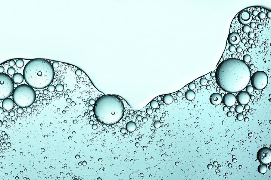 This image shows a close-up view of bubbles suspended in a liquid, likely oil droplets in water or a similar mixture. The bubbles vary in size and are clustered together, creating an interesting pattern against a light blue background. The image highlights the surface tension and interaction between the two immiscible liquids.
