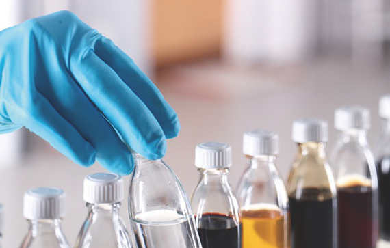The image shows a gloved hand handling a small glass vial or bottle in a lab-like setting, with several other sample bottles lined up in the background.