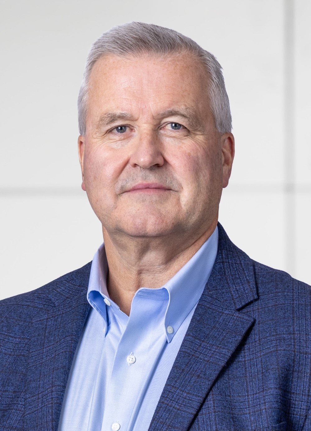 Middle-aged man wearing a blue checkered blazer and light blue dress shirt against a plain light background.