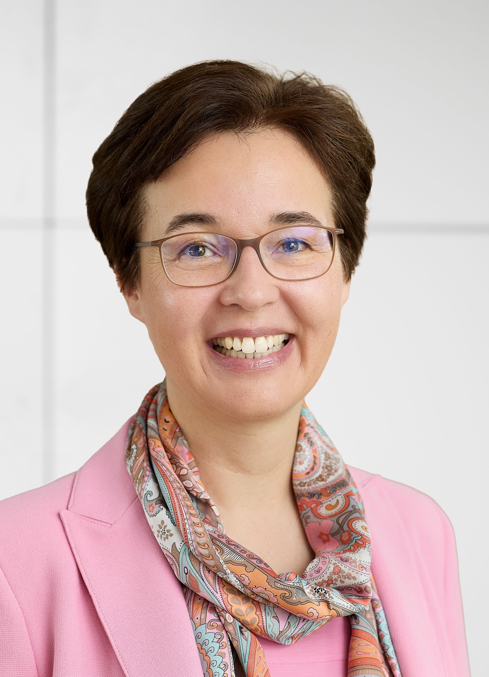 The person in the image is wearing a light pink blazer and a colorful scarf with a paisley pattern that includes shades of orange, pink, blue, and white. The background is plain and light-colored, possibly white or light gray. The person's face is blurred, so no facial details can be described. The hairstyle is short and brown. If you need any specific information or analysis, please let me know!