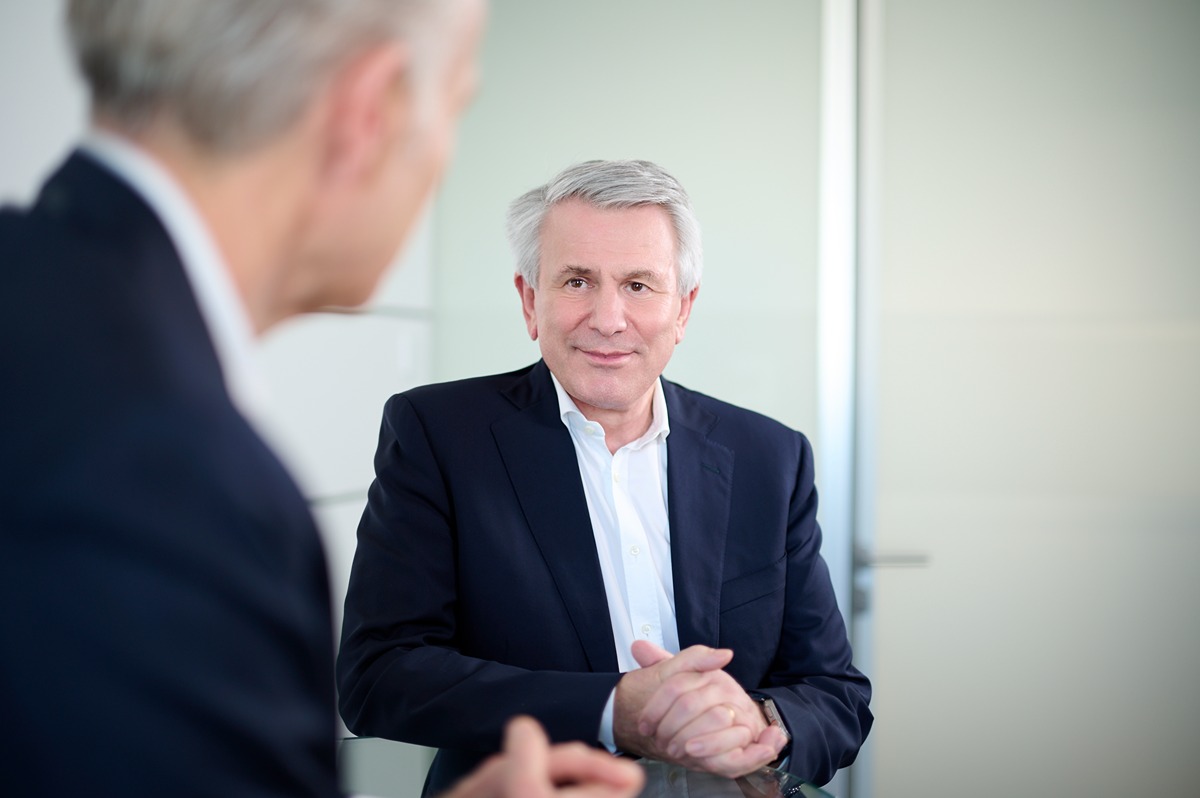 The image shows two older men in a professional setting, likely an office. Both men have gray or white hair and are wearing dark suits with white shirts. One man is facing the camera with his hands clasped in front of him on a glass table, while the other man is seen from behind, slightly out of focus. The background is minimalistic with light-colored walls and a door. The overall atmosphere suggests a formal meeting or discussion.