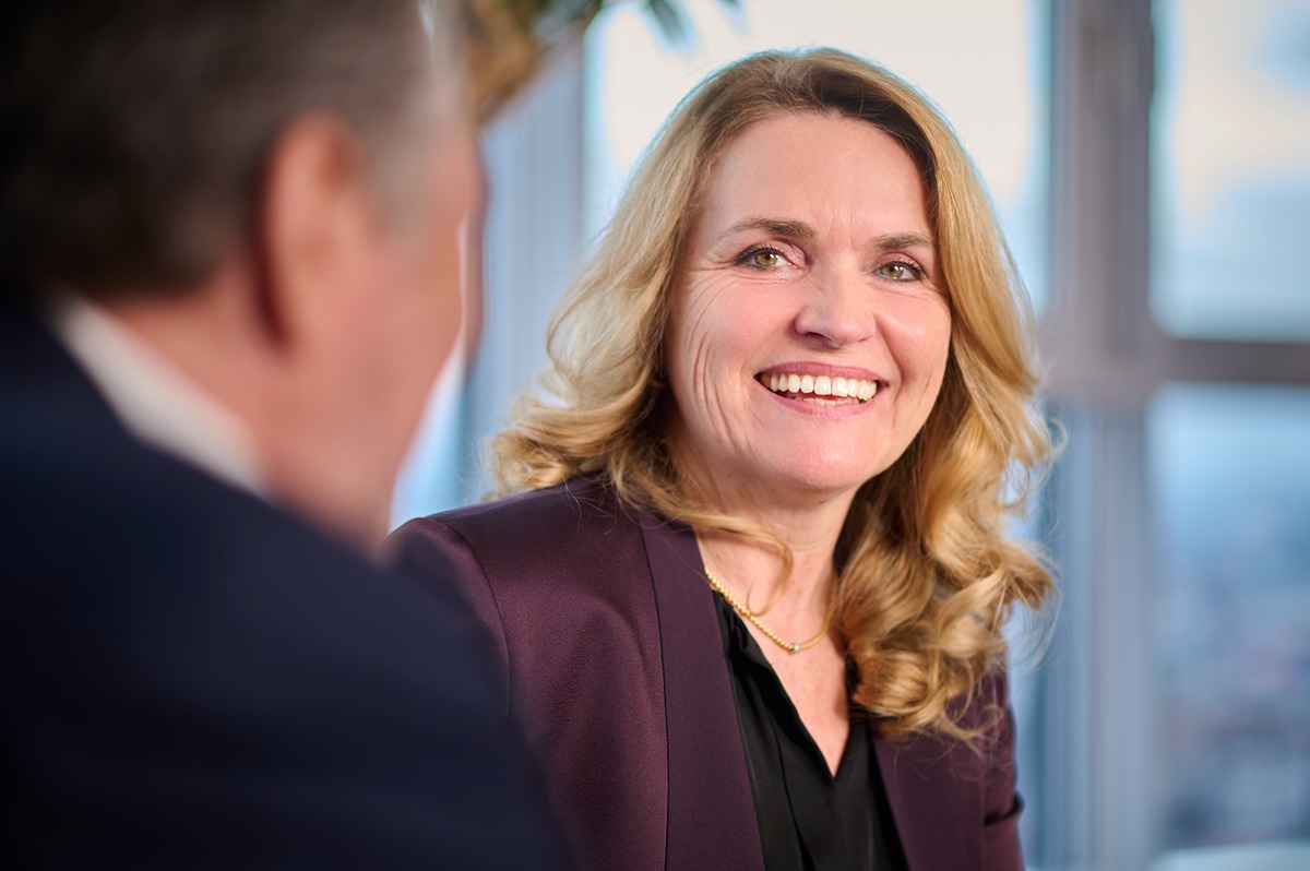 Two business professionals engaged in a conversation in a modern office setting with large windows and natural light.
