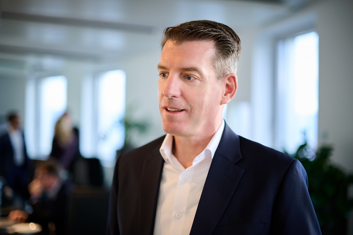 The person in the image is wearing a dark navy blue suit jacket over a white dress shirt. The setting appears to be a professional or office environment, with several other people in the background, some of whom are blurred. The lighting is bright, coming from large windows, suggesting a modern office space. The person's hair is neatly styled, combed back, and appears to be light brown or dark blonde.