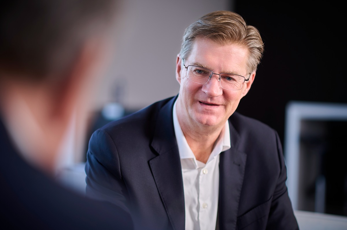 Two men in business attire engaged in a conversation in an indoor setting with a blurred background.