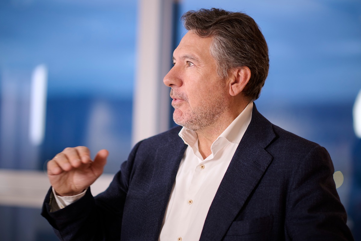 The person in the image is wearing a dark navy blue blazer over a white dress shirt. The shirt is buttoned up, and the blazer appears to be formal or semi-formal. The individual has short, dark brown hair. The background is blurred but suggests an indoor setting with large windows, possibly an office or a high-rise building with a view of the outside. The person is gesturing with their left hand, which is raised to about chest level.