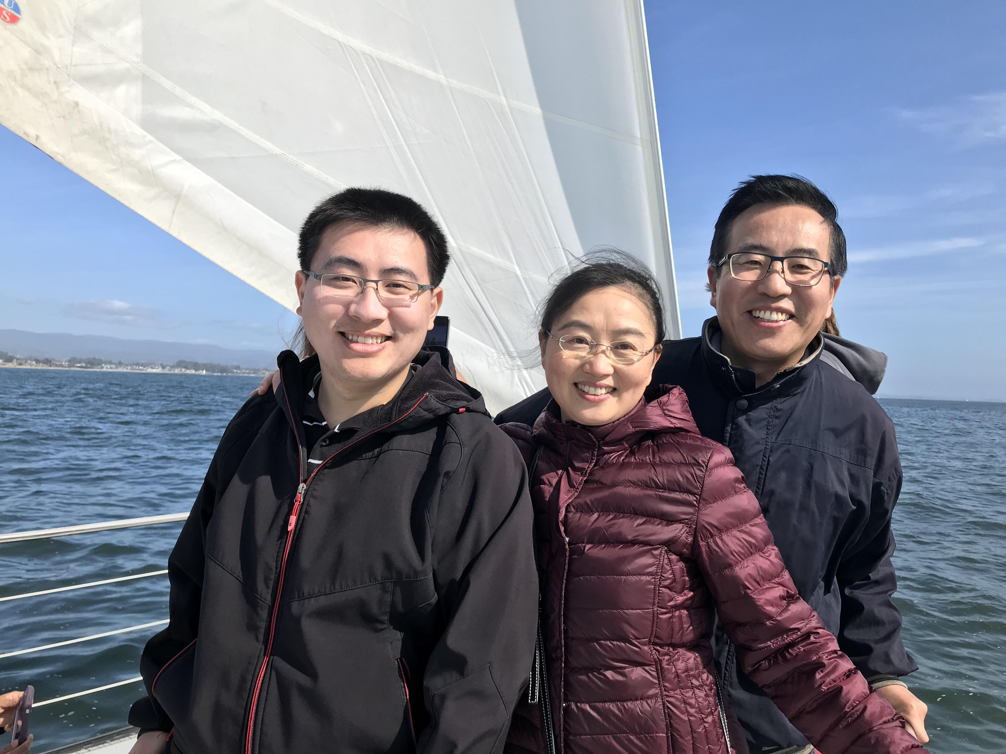 The image shows three people standing on a sailboat. They are dressed in jackets, suggesting it might be a cool or windy day. The sail of the boat is visible behind them, and the water and a distant shoreline can be seen in the background. The sky is clear with some light clouds. The overall setting appears to be a pleasant day for sailing.
