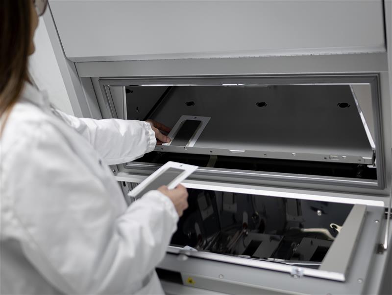 The image shows a person wearing a white lab coat working with a piece of laboratory equipment. The person is holding a rectangular object and appears to be placing or removing a similar object inside the machine. This setup suggests a scientific or medical laboratory environment, possibly involving sample analysis or preparation.