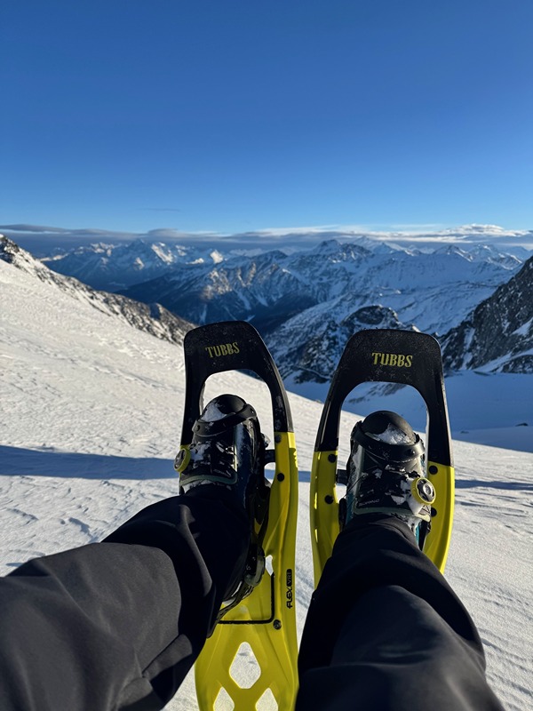 This image shows a person wearing snowshoes on a snowy mountain slope. The snowshoes are black and yellow and branded "TUBBS." The person is wearing black pants and black boots. In the background, there are snow-covered mountains under a clear blue sky. The scene suggests a winter outdoor activity, likely snowshoeing or hiking in a mountainous area.