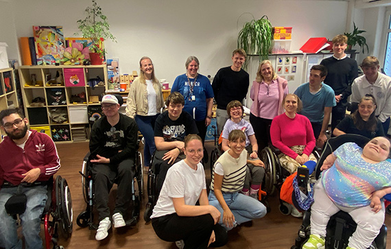 This image shows a group of people, some of whom are in wheelchairs, gathered together in what appears to be a community or activity room. The room has shelves with various items, including toys and books, and some plants. The group seems to be diverse in terms of age and gender, and they are posing for a group photo. The atmosphere looks friendly and inclusive.