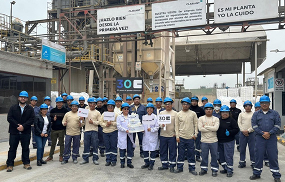In the image, there is a group of industrial workers standing together in front of a large industrial facility. Most of the workers are wearing blue helmets and beige or blue uniforms, while two individuals in the center are dressed in white lab coats and helmets, holding signs and a cake. The signs read "30 E" and "CLARIANT," indicating a celebration, likely the 30th anniversary of the company or plant.

Above the group, there are three large banners with Spanish text:
1. "¡HAZLO BIEN DESDE LA PRIMERA VEZ!" (Do it right the first time!)
2. "NUESTRA VISIÓN: Ser empresa líder mundial en especialidades químicas, destacando por encima del promedio, ofreciendo valor sustentable para nuestros grupos de interés." (Our vision: To be a world-leading company in chemical specialties, standing out above the average, offering sustainable value for our stakeholders.)
3. "ES MI PLANTA YO LA CUIDO" (It's my plant, I take care of it.)

The setting appears to be a chemical or industrial plant, and the group is likely celebrating a milestone related to the company Clariant.