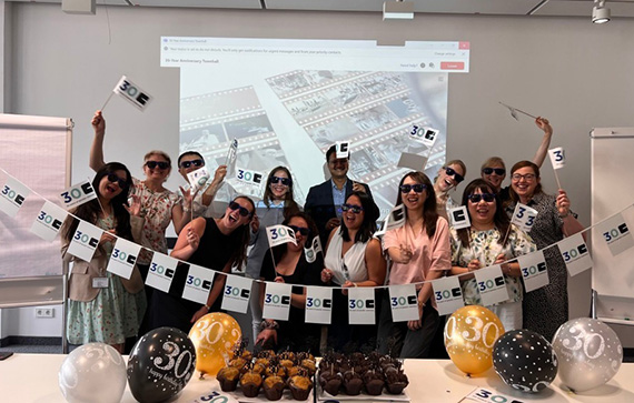 This image shows a group of people celebrating a 30th birthday or anniversary event. They are holding small flags and a banner that say "30" with a black and white design. There are balloons in black, gold, and white colors with the number "30" printed on them. On the table in front of the group, there are trays of chocolate-covered treats or cake pops. The background features a projection screen displaying images of film strips, suggesting a theme related to movies or memories. The overall atmosphere appears festive and joyful.