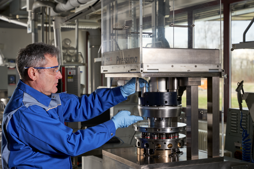 The image shows a person in a blue lab/work uniform and gloves operating or adjusting a large industrial machine in a workshop or lab setting. The machine appears to be a precision press or testing apparatus with a transparent protective enclosure.