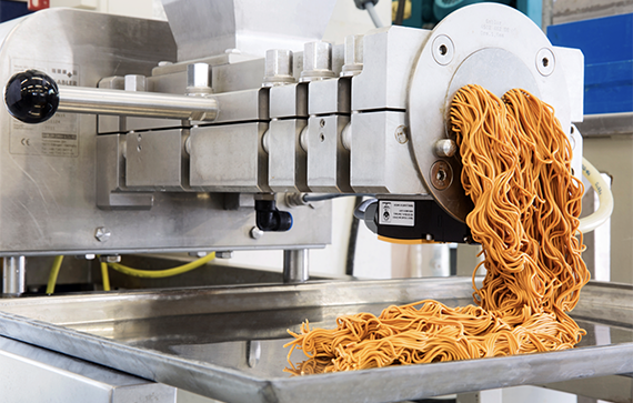 The image shows an industrial food-processing machine extruding a mass of orange noodles or pasta into a metal tray.