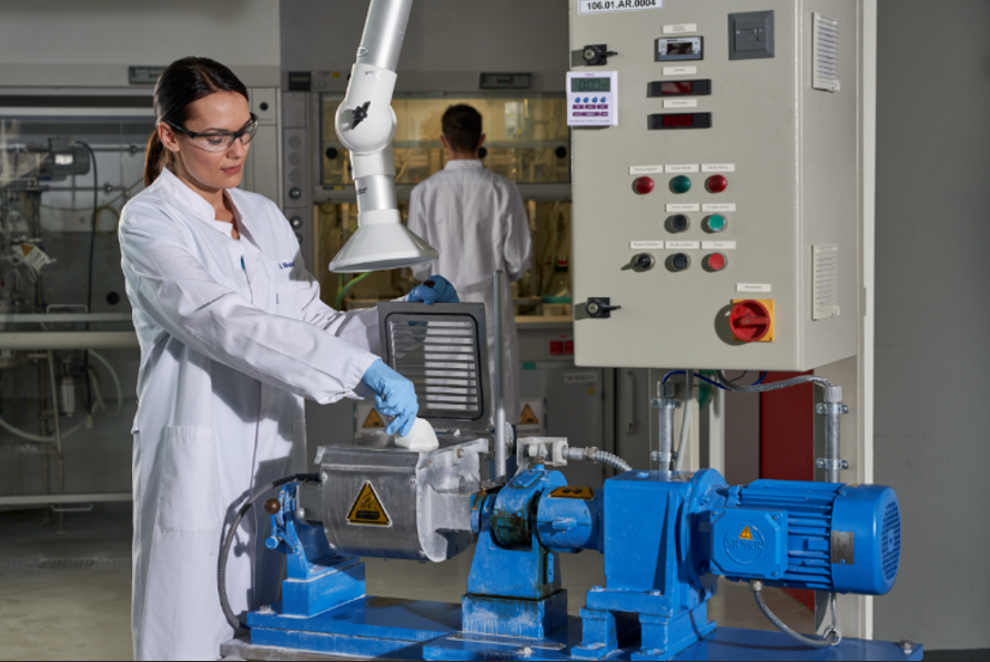 The image shows a laboratory or industrial testing setup. A person in a white lab coat and blue gloves is operating a blue machine with a control panel nearby. Another person in a lab coat is visible in the background. The scene appears to be in a controlled lab environment with machinery and safety equipment.
