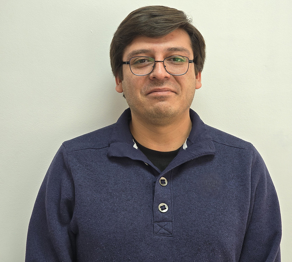 The person in the image is wearing a dark blue pullover with a collar and two buttons. The background is a plain light-colored wall. The person's face is blurred, so no facial details are visible. The hair is brown and neatly combed. The overall appearance is casual.