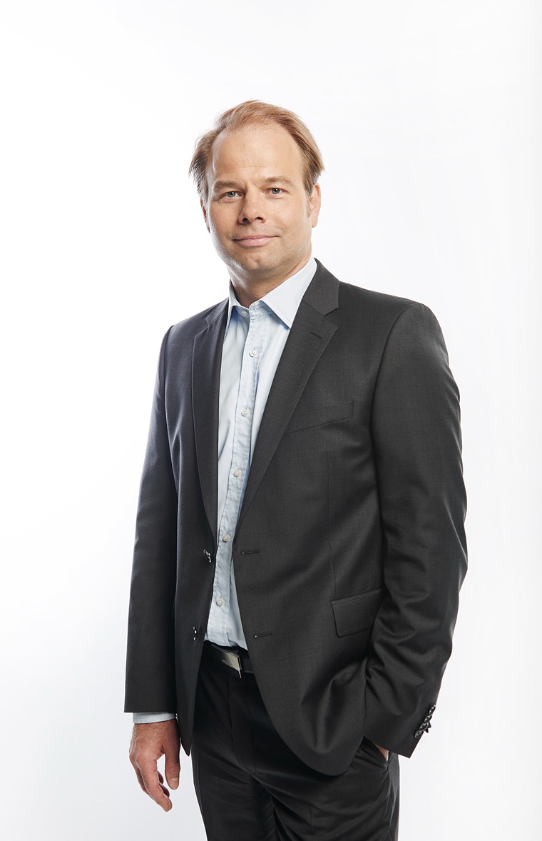 The person in the image is wearing a formal outfit consisting of a dark gray suit jacket, a light blue dress shirt, and black trousers. The shirt is buttoned up but without a tie. The individual has light brown or blonde hair and is standing with one hand in their pocket and the other hand relaxed by their side. The background is plain white, giving the image a professional and clean appearance.