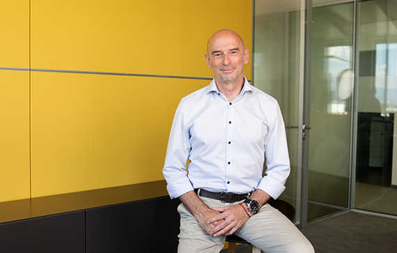 The image shows a man sitting on a stool in an office setting. He is wearing a light blue button-up shirt with the sleeves rolled up and light-colored pants. He has a watch on his left wrist and a belt. The background features a yellow wall panel on the left and glass office partitions on the right. The man's face is blurred for privacy.
