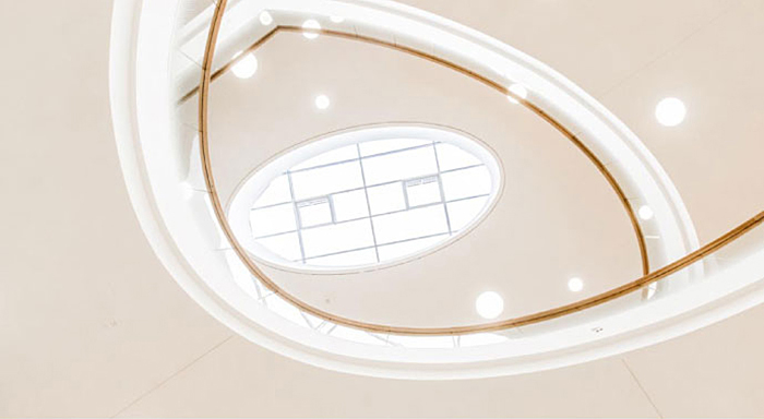 The image shows a bright, modern ceiling with a curved oval skylight or light well in the center, surrounded by recessed circular lights and a gold-toned trim or railing.